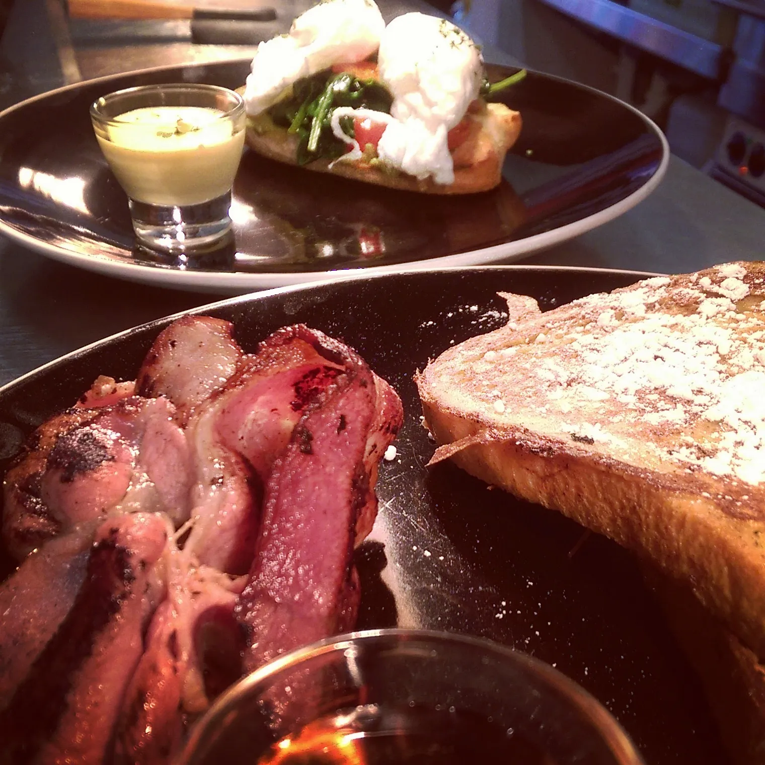 Two breakfast plates with bacon and French toast in foreground, poached eggs in background