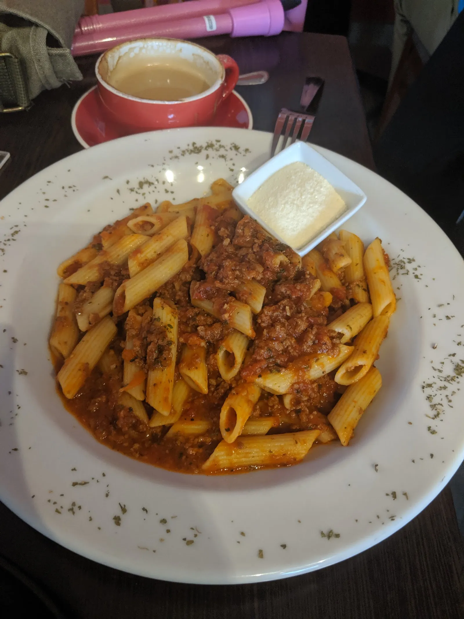Penne bolognese with parmesan ramekin on white plate, coffee cup in background