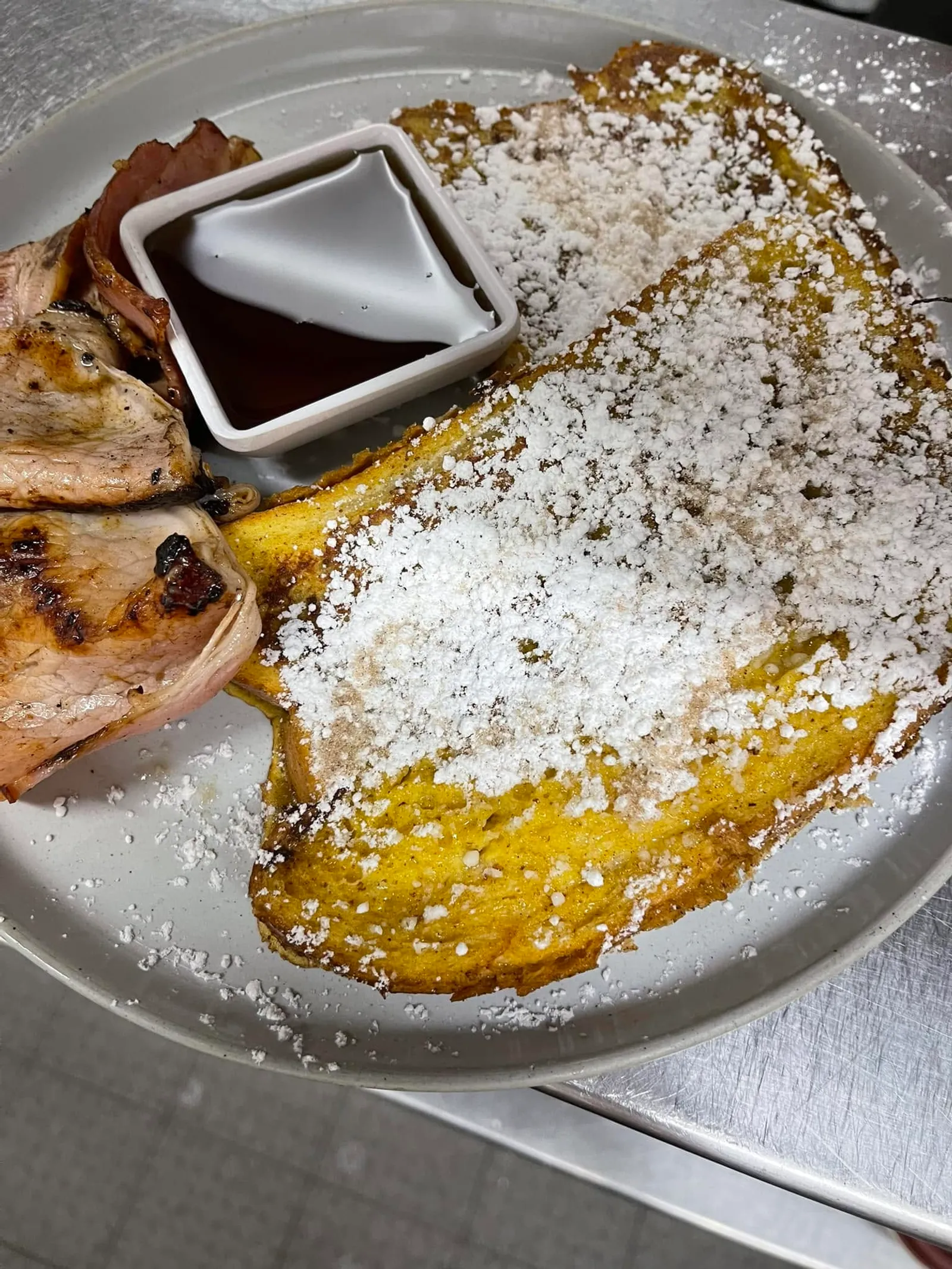 French toast with icing sugar, bacon and maple syrup on white plate